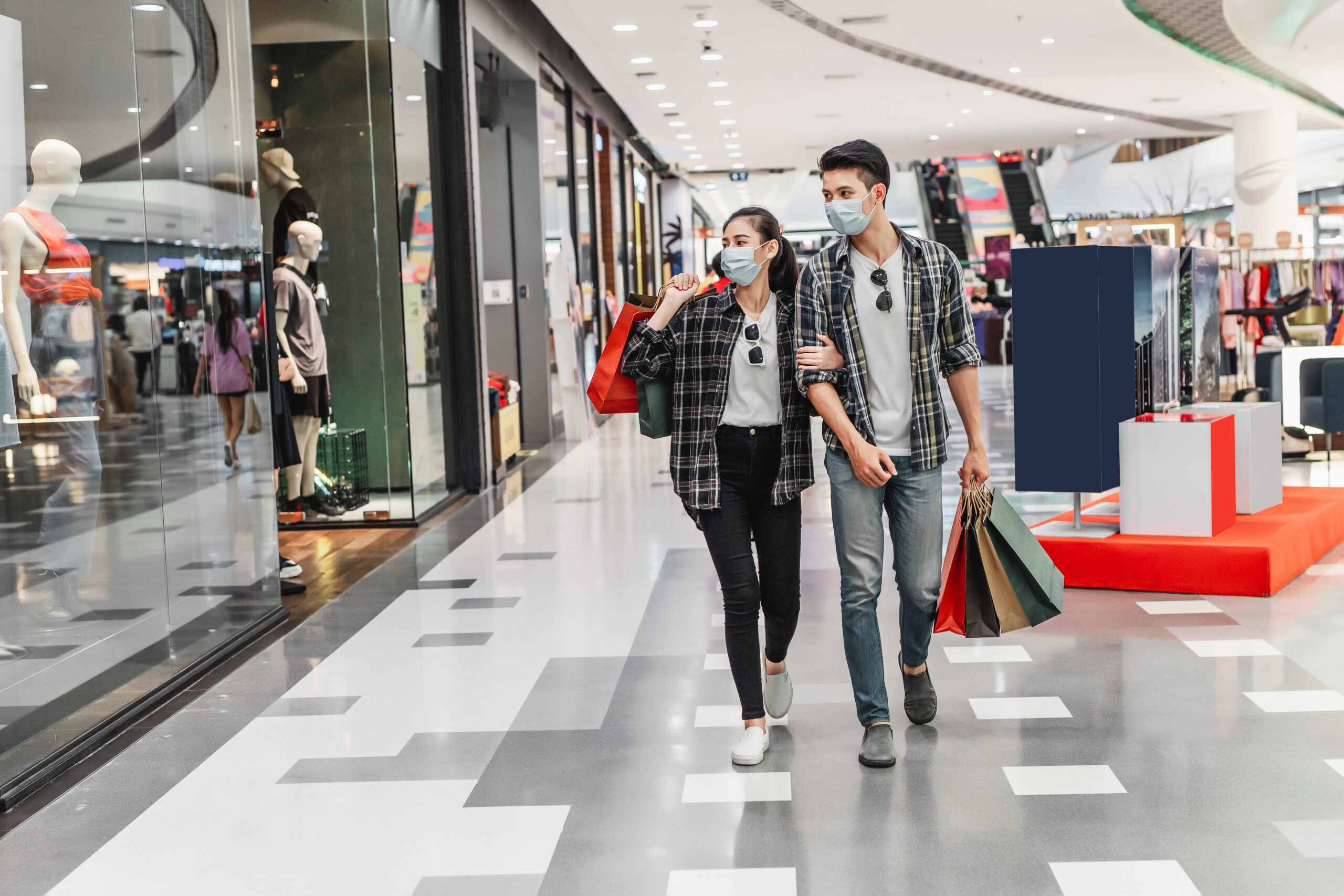 Young couple in protection mask holding multiple paper shopping bag walking in the corridor of large shopping mall, New normal lifestyle and shopping concept